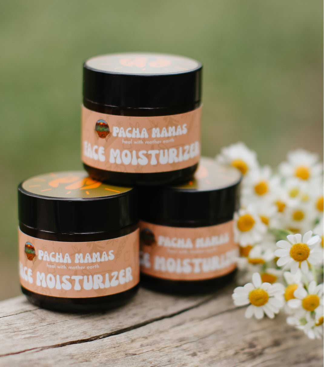 Three jars of Pacha Mamas Face Moisturizer on a wooden surface with flowers in the background.