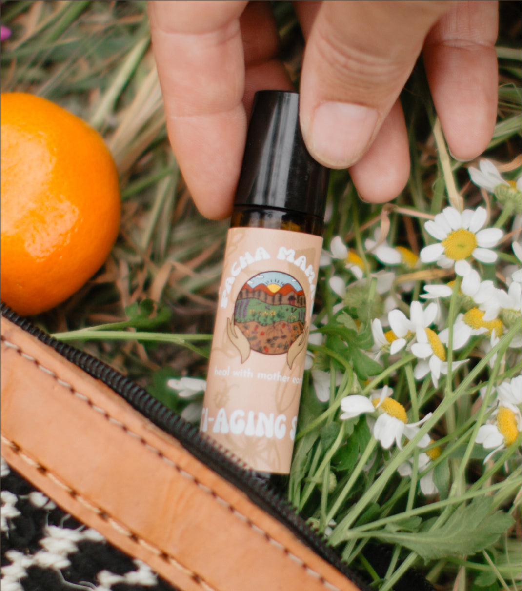 Roll-on bottle with a label featuring an illustration of a landscape, held by a hand among flowers and an orange.